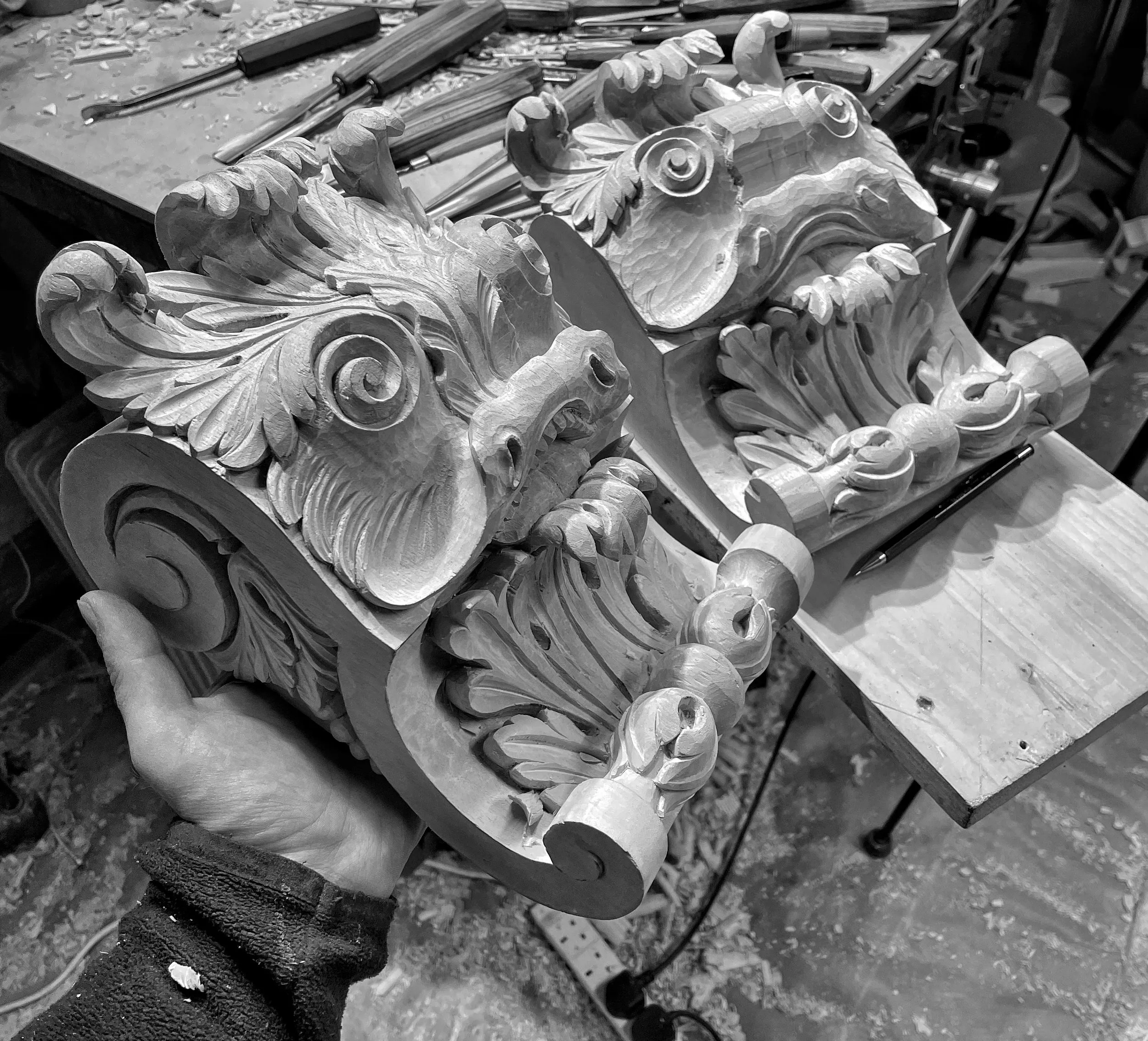 A pair of hand-carved acanthus scroll brackets on a workbench, surrounded by carving gouges